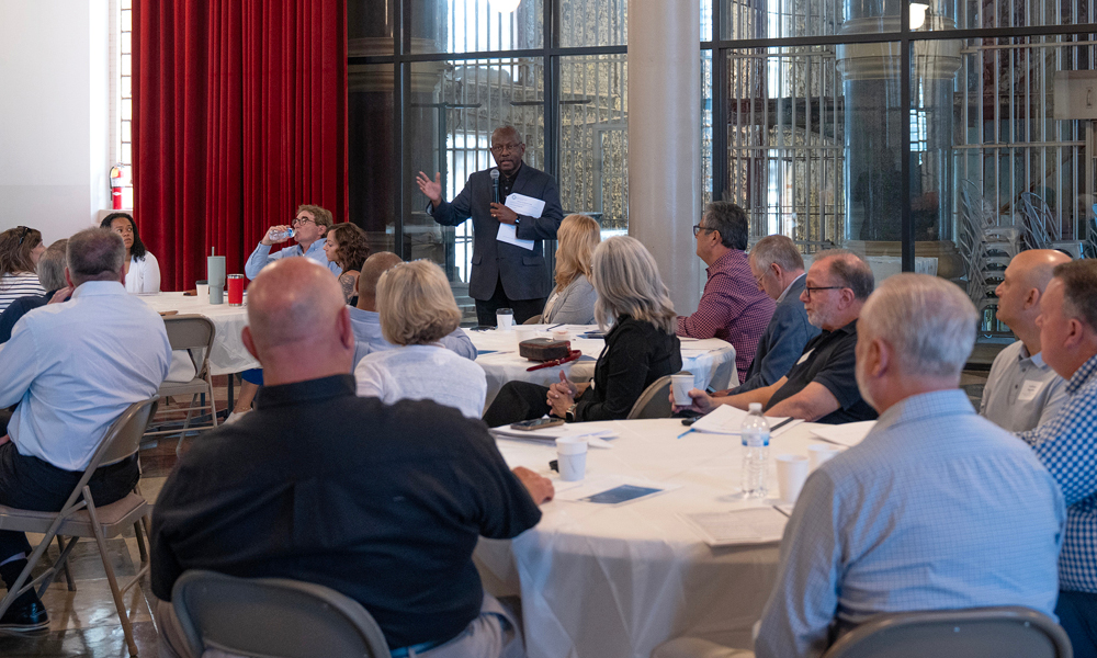 A woman wearing black pants and a green, short-sleeve shirt passes out papers to a roomful of men and women seated at long tables.